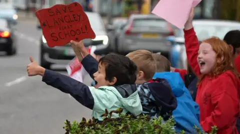 A group of young boys facing cars on the road with placards. One boy is holding up a handmade sign which reads Save Langley First School in one hand and holding his thumb up with his other.