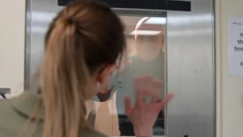 A woman with a blonde ponytail waves from behind through a glass panel of a steel door, through which a bald man in a black polo neck can be seen laughing as he waves back.