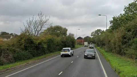 Google Cars are pictured driving on a road in both direction. The road is lined by a grass verge and hedges. Buildings can be seen in the distance as well as lamp posts.