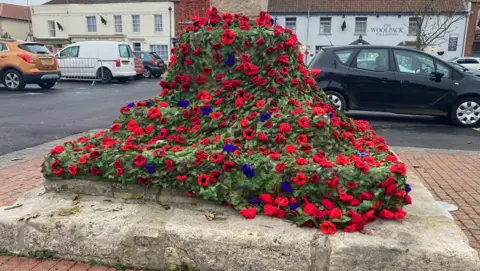 The poppies are attached to a green netting, and there are also blue flowers on there of some description.  The Buttercross is on a stone plinth on three levels and the netting is draped all around the bottom.