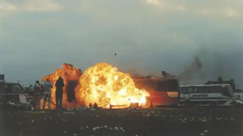 Only Fools and Horses Appreciation Society A coach surrounded by a large explosion, with flames and smoke rising into the air in an outdoor setting.