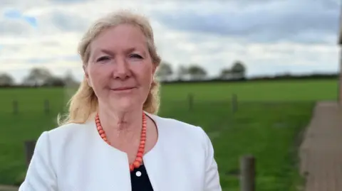A middle-aged woman with blonde shoulder length hair swept back behind her ears. She is smiling at the camera and wearing a white collarless jacket a black top and orange beaded necklace. In the blurred background is a grass field and a line of trees and a hedge in the distance.