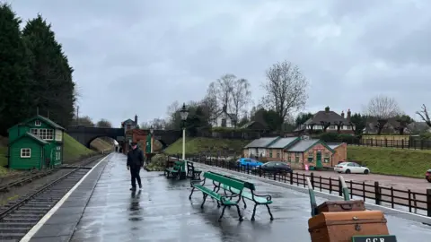 Station platform with benches and one man, and old fashioned luggage on a trolley, with tea rooms on the right