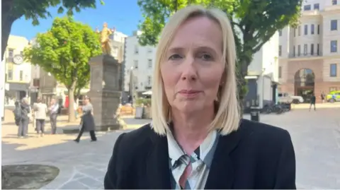 BBC Deputy Catherine Curtis is pictured in Jersey's Royal Square. She has blonde hair, is wearing a blazer and a green shirt. 