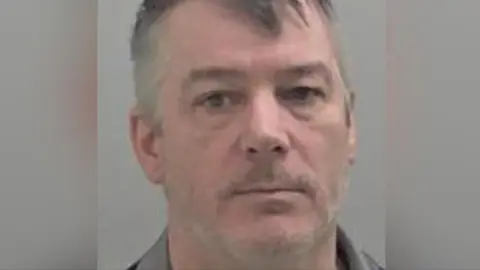 Michael Morris seen in a police mugshot. He has a greying beard and short hair and is looking at the camera.