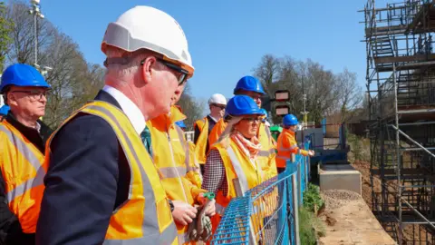 West Midlands Combined Authority Parker, the person nearest to the camera, is wearing a hard hat and hi-vis jacket, along with half a dozen others on the left and middle of the photo. They are looking towards a building site on the right.