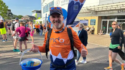 Katy Lambert/BBC A man in an orange MND association t shirt. He has a flag on his back and a collection bucket which, today, contains bottles of water instead of coins
