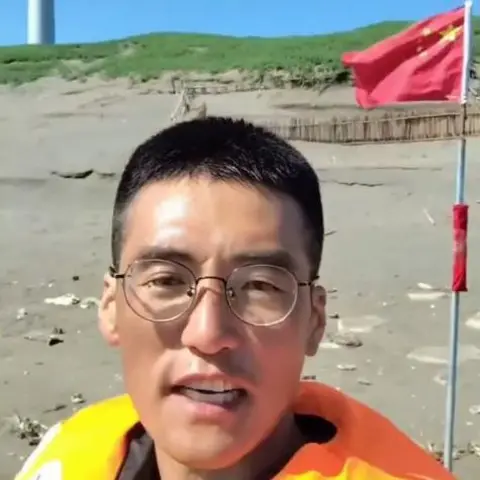 Shandong Kaige Chinese influencer Shandong Kaige addresses the camera wearing what appears to be an orange lifesaver vest, standing on a beach, with a Chinese flag planted in the sand behind him.