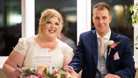 Rose Studio Photography Amy Campbell on the left wearing a lace wedding dress, curly hair and a pendant. She has curly blonde hair tied up. She holds a pen. Her husband is on the right and is wearing a navy blue suit, silver vest and pink tie, with a pink and white rose attached to him. They are both smiling and holding hands. 