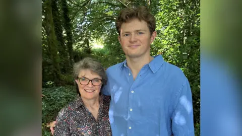 The image shows two people standing in a woodland setting, with trees and greenery around them. On the left is a man wearing a blue shirt, and on the right is a woman with grey hair and glasses.