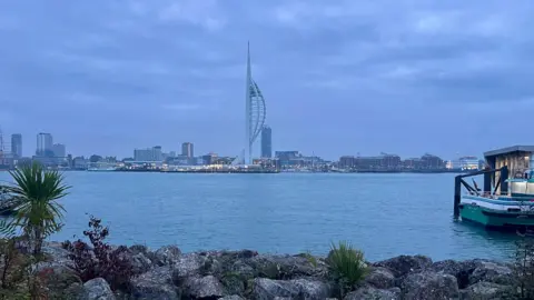 tonyswinton Portsmouth's skyline seen behind a large body of water and under grey clouds, with the Spinnaker Tower in the centre