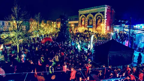 Kettering Town Council A photo of Kettering town centre with buildings illuminated in lots of different coloured lights. Large crowds are gathered around a large Christmas tree.