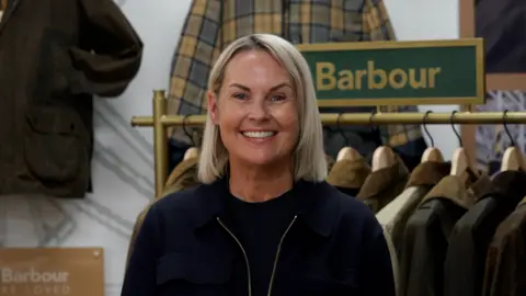 Hannah Karpel/BBC Woman standing in the foreground, wearing a dark jacket and top, facing the camera inside a clothing retail display. Behind the person is a rail of waxed jackets on wooden hangers and a green sign reading “Barbour”. Additional jackets hang on the wall, including a checked garment. A plaque on the left displays the text “Barbour Re-Loved”. The setting appears to be a branded clothing shop or exhibition area.