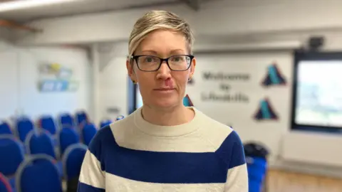 A woman with short blonde hair wearing a blue and white jumper and black glasses. The room behind her is blurred, showing blue chairs in rows.