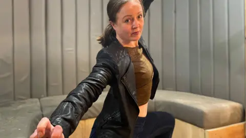 Simon Thake A woman in a black leather jacket kneels with her hand extended in a fight pose.