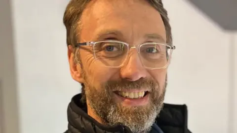 A man with brown hair and a beard with silver-framed glasses and a high black collar