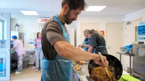 Man in a kitchen holding a frying pan and pouring stir fry into box