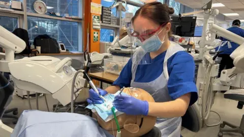 Woman with a model head and dental equipment. She is wearing a blue T-shirt and she has goggle and black hair. Lots of medical equipment is behind her