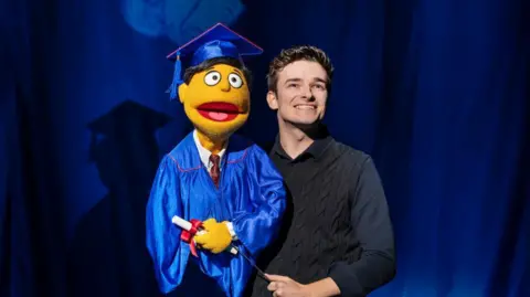 Matt Crockett A man stands on stage beside a yellow puppet character dressed in a blue graduation gown and cap, holding a diploma.
