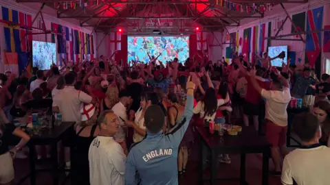 Crowds of people have their arms raised towards a screen, watching a football match.