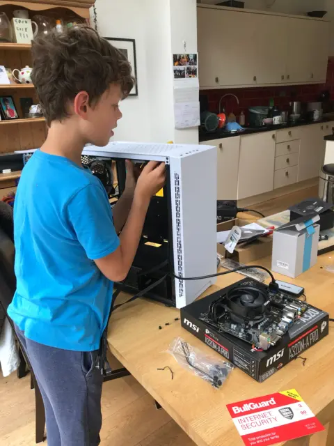 family handout Toby, a seven-year-old boy, is building computer. He is wearing a blue T-shirt and is holding a metal object inside a white metal case. In the background is a bookcase full of ornaments, books and photographs.