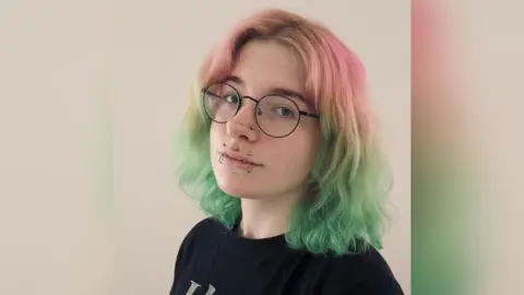 Portrait photo of young woman with pink and green hair, round glasses and a number of lip piercings.