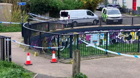 Police tape cordoning off a bridge over an underpass on Honeybourne Way, Gloucestershire. A plain white van and a police van are parked on the road.