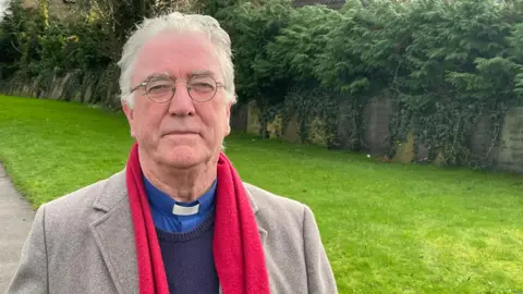 A vicar stands in front of a green lawn in the grounds of a village church. He has white hair and is wearing glasses, a blue shirt with white dog collar, a blue pullover, taupe-grey jacket and red scarf.