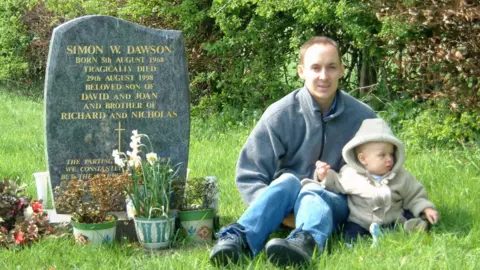 Nick Dawson A man holding a young child sat next to a grave. The grave has a black stone with gold writing reading: "SIMON W. DAWSON".