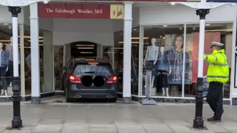 A stationary black Mercedes car sits in the doorway of the Edinburgh Woollen Mill shop on Lord Street in Southport. A police officer in a yellow high-vis jacket stands nearby.