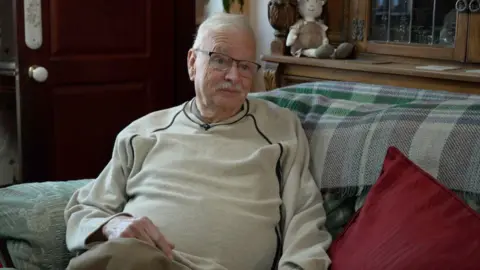 BBC/Ed Young An elderly man sits on a sofa. He is wearing a creme jumper and glasses.