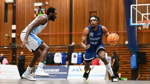 A person in a blue basketball kit drilling a basketball towards a person in a white basketball kit. The blue kit says "Worthing Thunder" on the front.