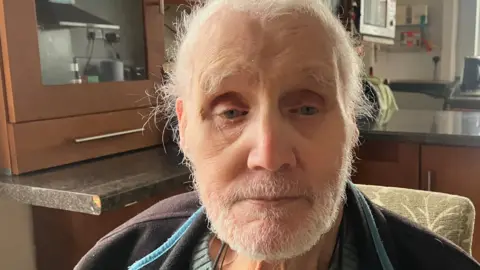 Richard Stead/BBC Roland Parkisson has white hair and beard and is photographed in his kitchen. He is wearing a dark fleece with sky blue zip and a bluey-green sweater.