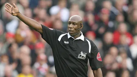 PA Media A man wearing a black referee's top with a Premier League logo on the chest. He is wearing a microphone and earpiece. He has one arm outstretched. In the background is a blurred view of fans in the stadium.