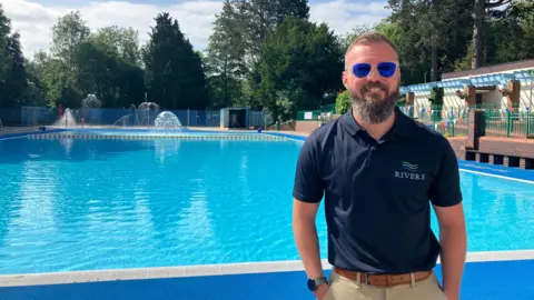 Seb Parker wearing sunglasses, a navy blue polo top and beige trousers stands beside a large outdoor pool. The water is bright blue and beyond the p