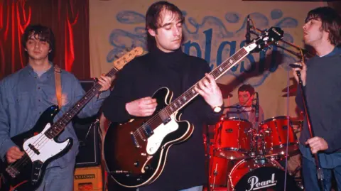 Getty Images Paul 'Guigsy' McGuigan, vestindo uma camisa jeans azul e jeans e segurando um baixo preto e branco, Paul 'Bonehead' Arthurs, vestindo um suéter preto com gola pólo e tocando uma guitarra marrom, Tony McCarroll, vestindo uma camisa jeans azul e tocando uma bateria vermelha, e Liam Gallagher, vestindo uma camisa azul e um suéter cinza-azulado, tocam em frente a uma placa marrom e azul que diz "Respingo" e uma cortina de veludo vermelho