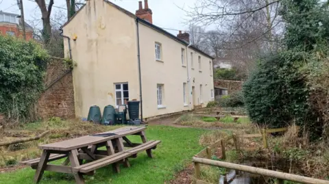 The Moorings, a two-story cream-colored house with a pitched roof and multiple chimneys. There are several small windows and doors visible. The surrounding area consists of a grassy garden with wooden picnic tables and benches in the foreground. A small stream runs beside the property, bordered by wooden railings. There are also green bushes and trees.