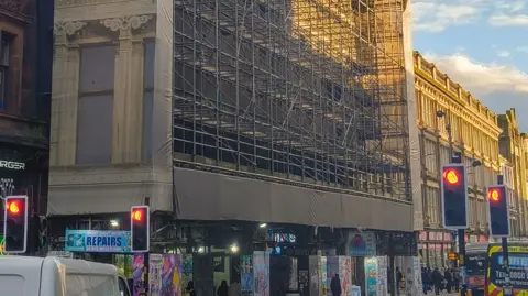 Glasgow City Council Scaffolding and canvas cover a wide building on a busy street. Traffic waits behind red lights and pedestrians walk on the pavement under the scaffold.