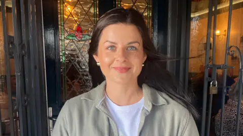 Dana has long black hair, metallic earrings and blue eyes. She is smiling at the camera.

She's wearing a grey shirt over a white t-shirt. 

She is standing in front of black doors and stained glass windows. 