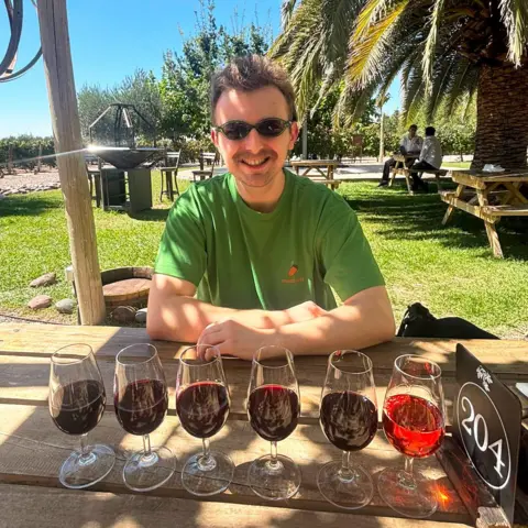 Dominic Lee Six glasses containing wine are lined up across a wooden table in the foreground. A person wearing sunglasses and a green T-shirt is seated behind the glasses. Picnic tables, palm trees, grass and vineyard rows are visible in the background.