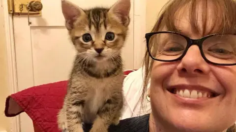 Lisa Burtenshaw is pictured with a small brown tabby kitten sitting on her shoulder. This is a close cropped shot in which only Lisa's face can be seen. She has brown hair and is wearing glasses with black frames. She and the kitten are looking directly at the camera and she is smiling widely.