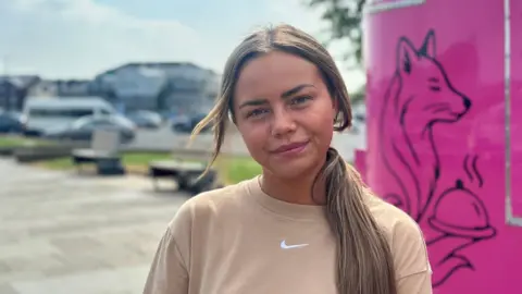 Rhiannon smiling at the camera. She is wearing a beige t-shirt with a white Nike tick on it. She has light brown hair that is tied in a low ponytail. 
