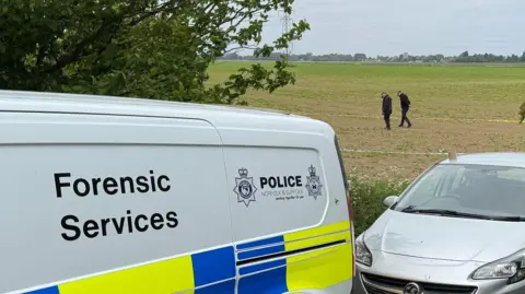 Steve Hubbard/BBC A forensic services van with Norfolk Police branding is parked in the foreground. Two police officers can be seen in a field behind examining the ground.