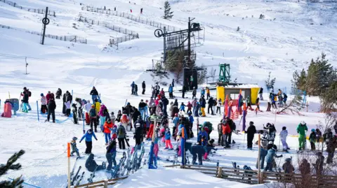 Crowds of snowsports enthusiasts in colourful ski wear at Cairngorm Mountain. It is a bright, sunny day.