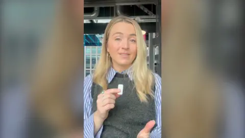 BBC Journalist Lucy Woodham wears a blue striped shirt with a grey pullover and looks at the camera. She holds a small white microphone with her right hand. 