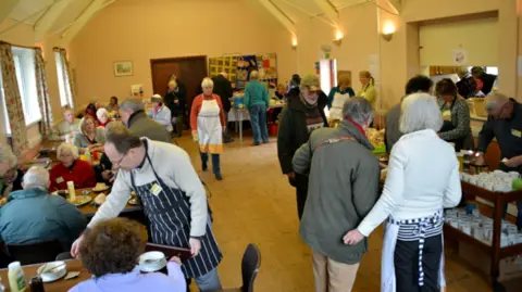 Robin Bennion Tables full of visitors enjoying refreshments in the village hall with lots of helpers in aprons walking about.