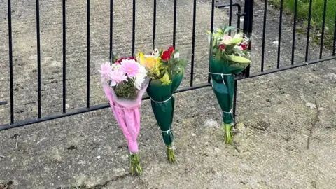 Three bunches of flowers placed on a driveway against a gate. 