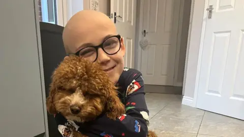 Fewster family Ezra with very short hair and glasses hugging a brown dog. He is wearing dark blue Christmas pyjamas and there are several doors of a domestic home behind him.