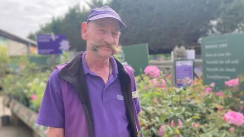 A man with a large handlebar moustache, wearing a purple uniform consisting of a baseball cap, polo shirt, and fleece bodywarmer. He is smiling and there are rows of flowers behind him in an outdoor area of the garden centre.