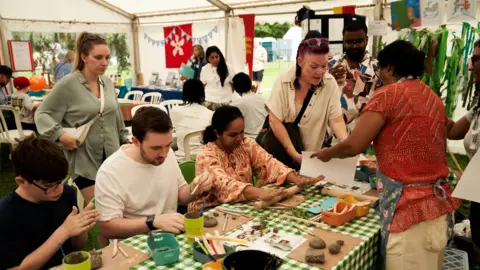 Andy Willsher/BCA A group of people inside a marquee, taking part in a crafting activity. A number of people are sat down, a few more are standing up. There are craft items in front of them and lots of chairs and tables in the room. 